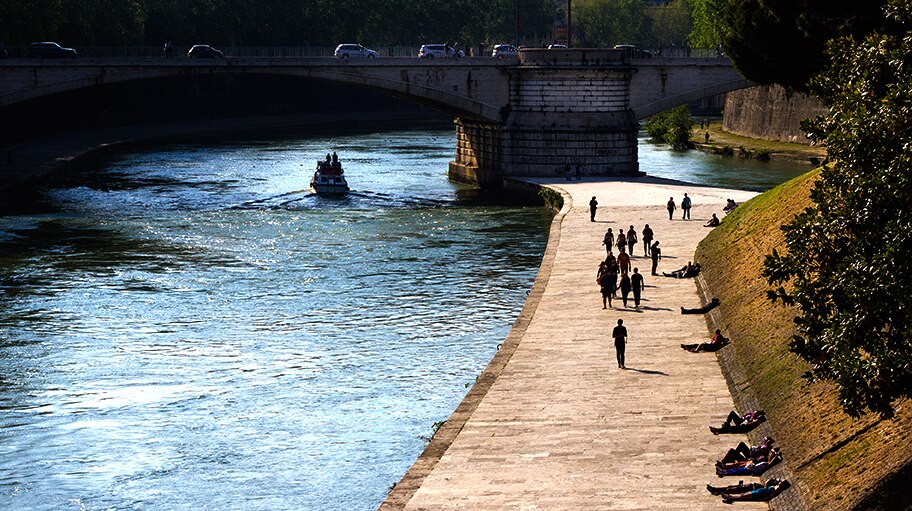 People Walking Next To A River Bridge In The Background