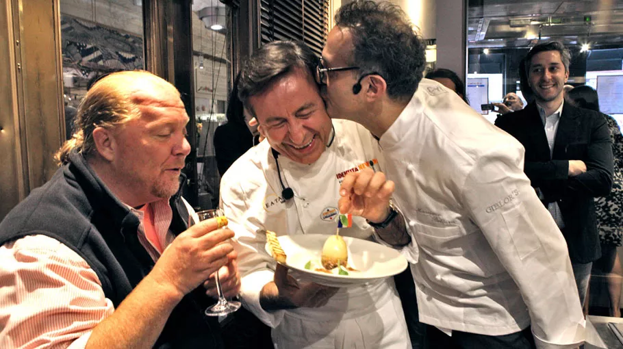 Two chefs laughing while holding a plate with a smiling man on the background