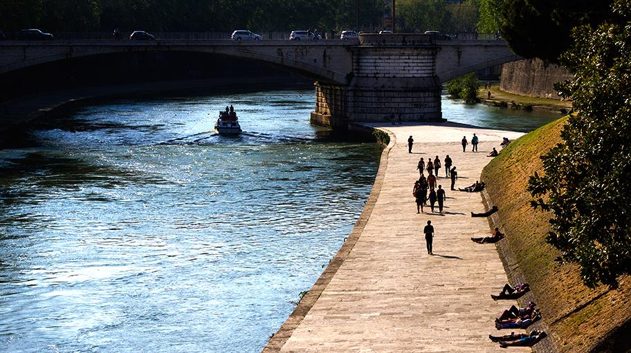 People Walking Next To A River Bridge In The Background