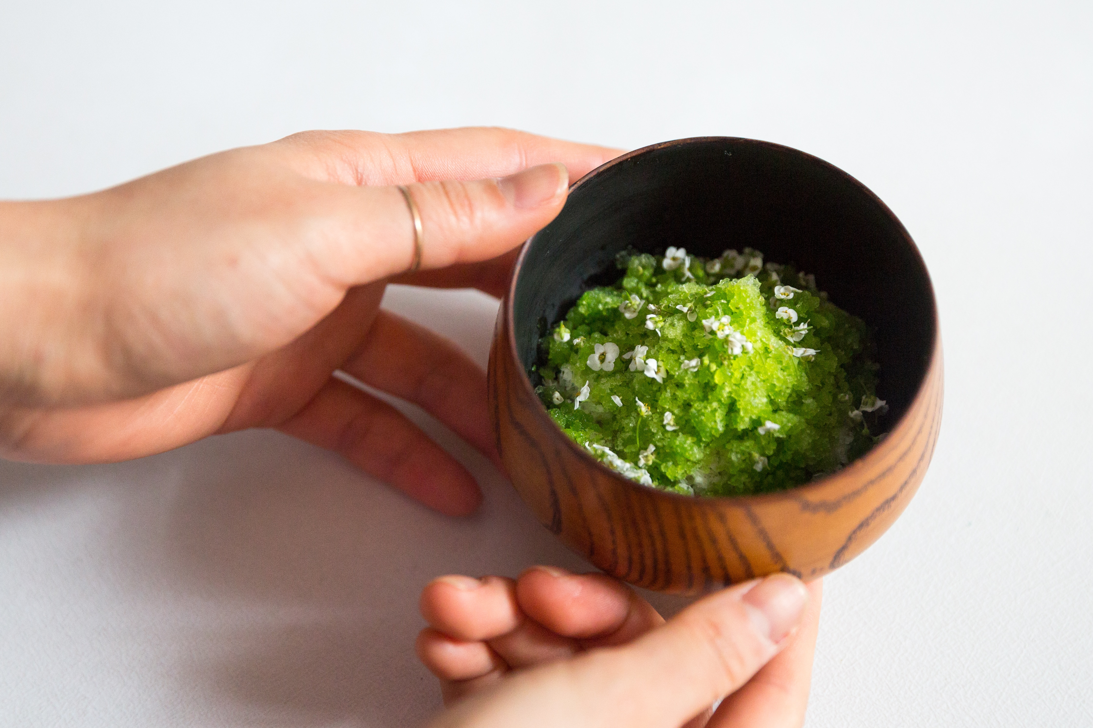 Green granita with white flowers in a wooden bowl, held by two hands.
