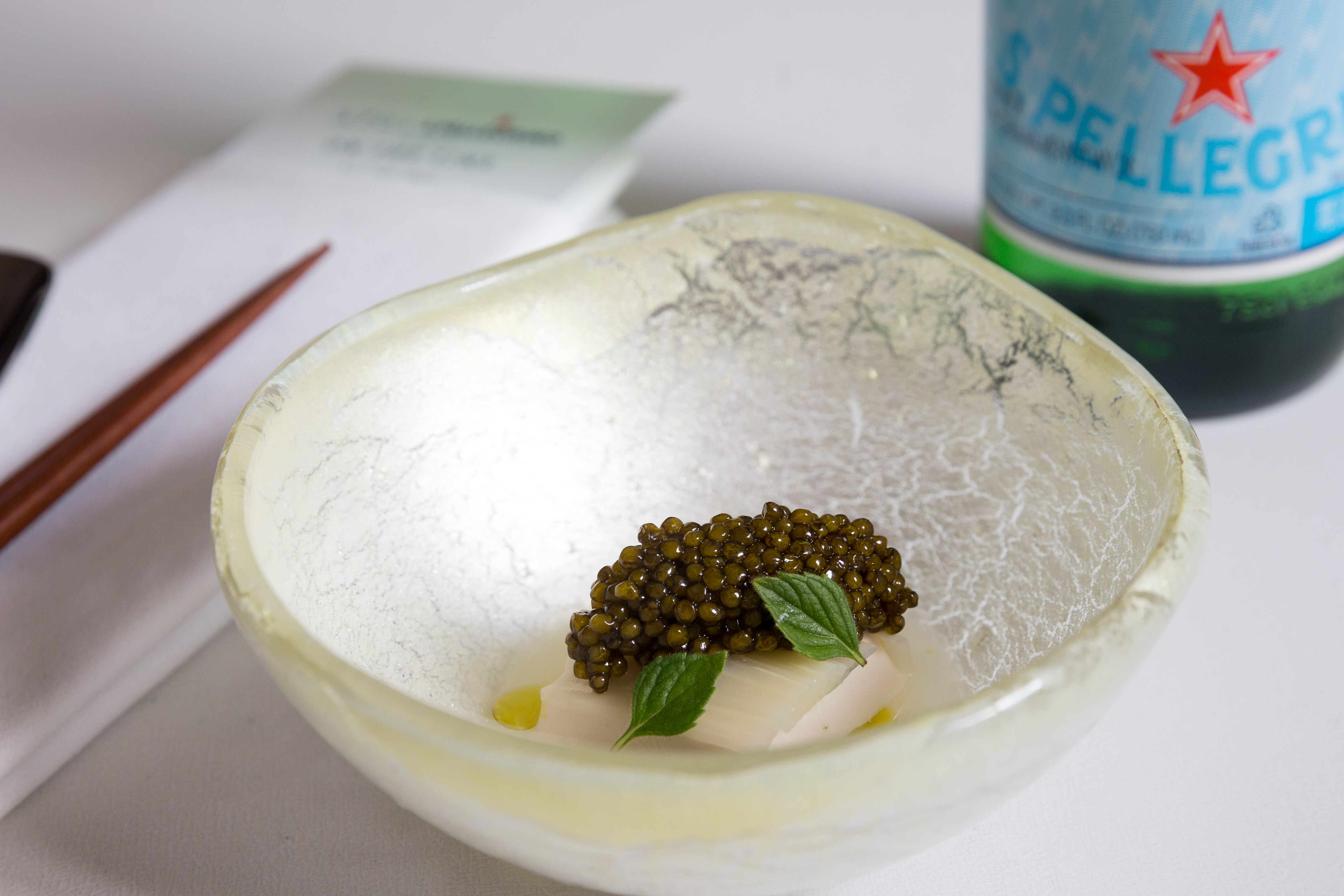 White bowl with hiramasa, caviar, and herbs, with a San Pellegrino bottle and chopsticks nearby.