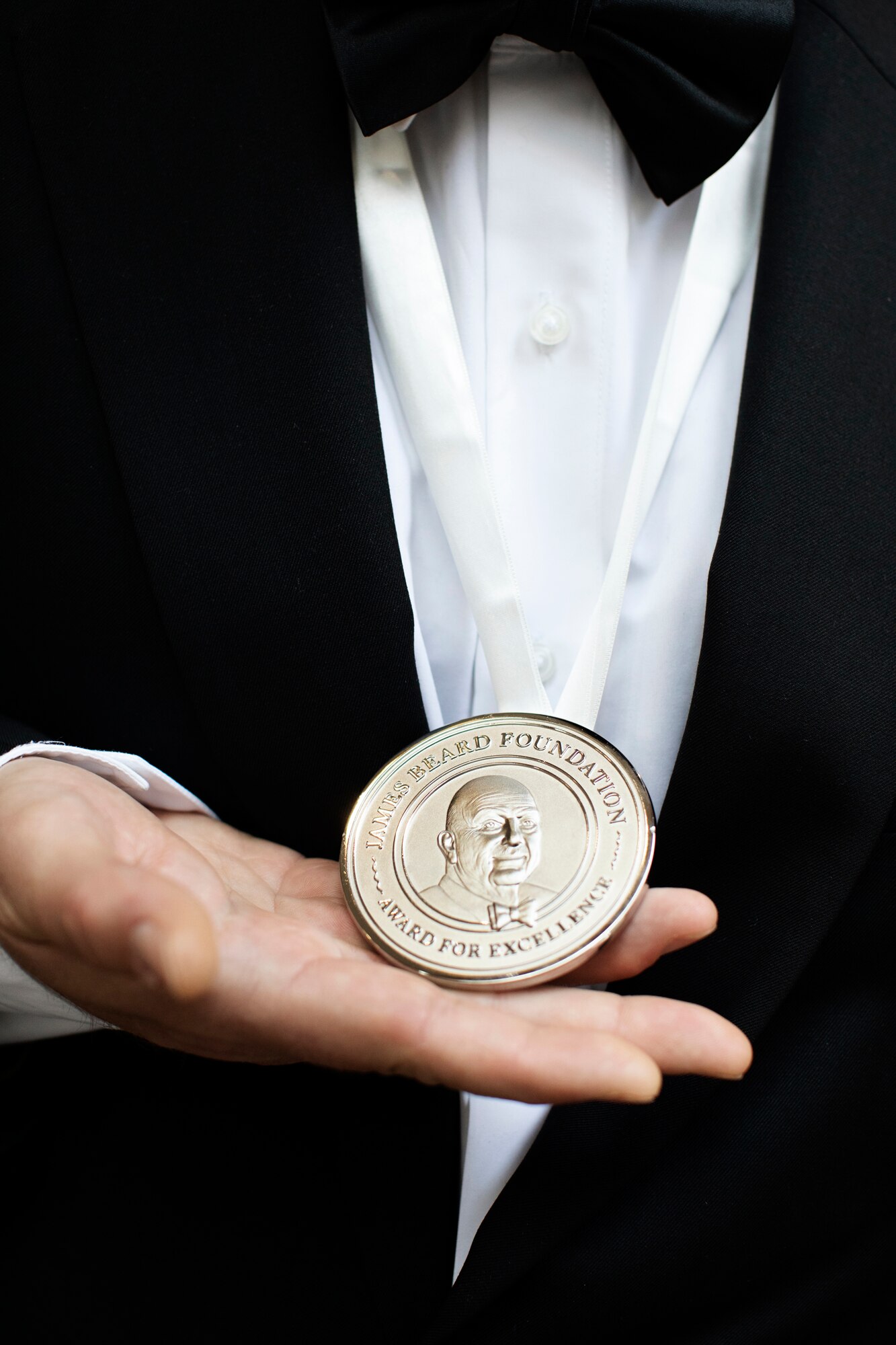 Close-up of a person holding a James Beard Foundation medal while dressed in a black tuxedo.