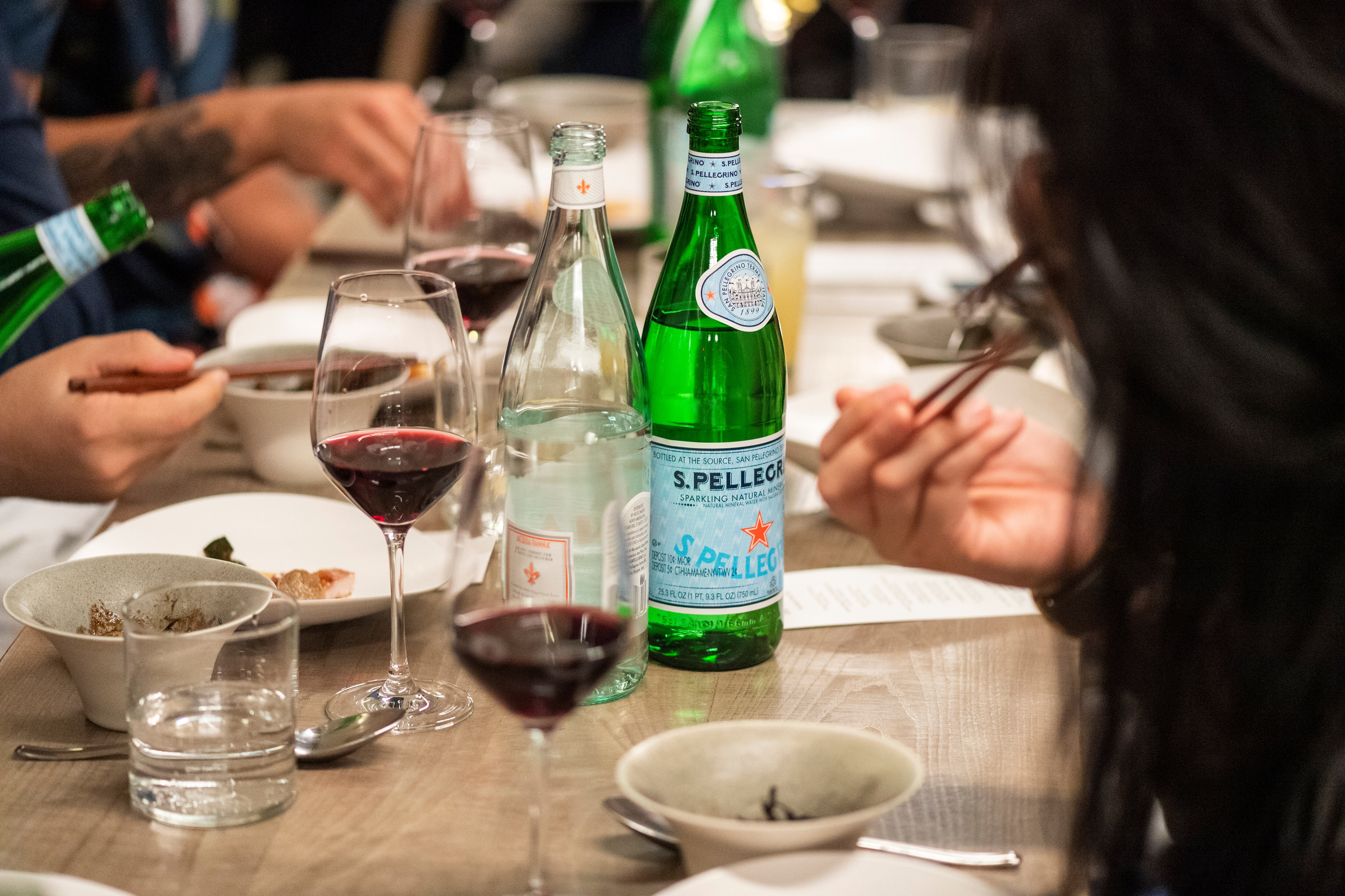 Group dining with San Pellegrino, wine, and food at a long table, diners using chopsticks.