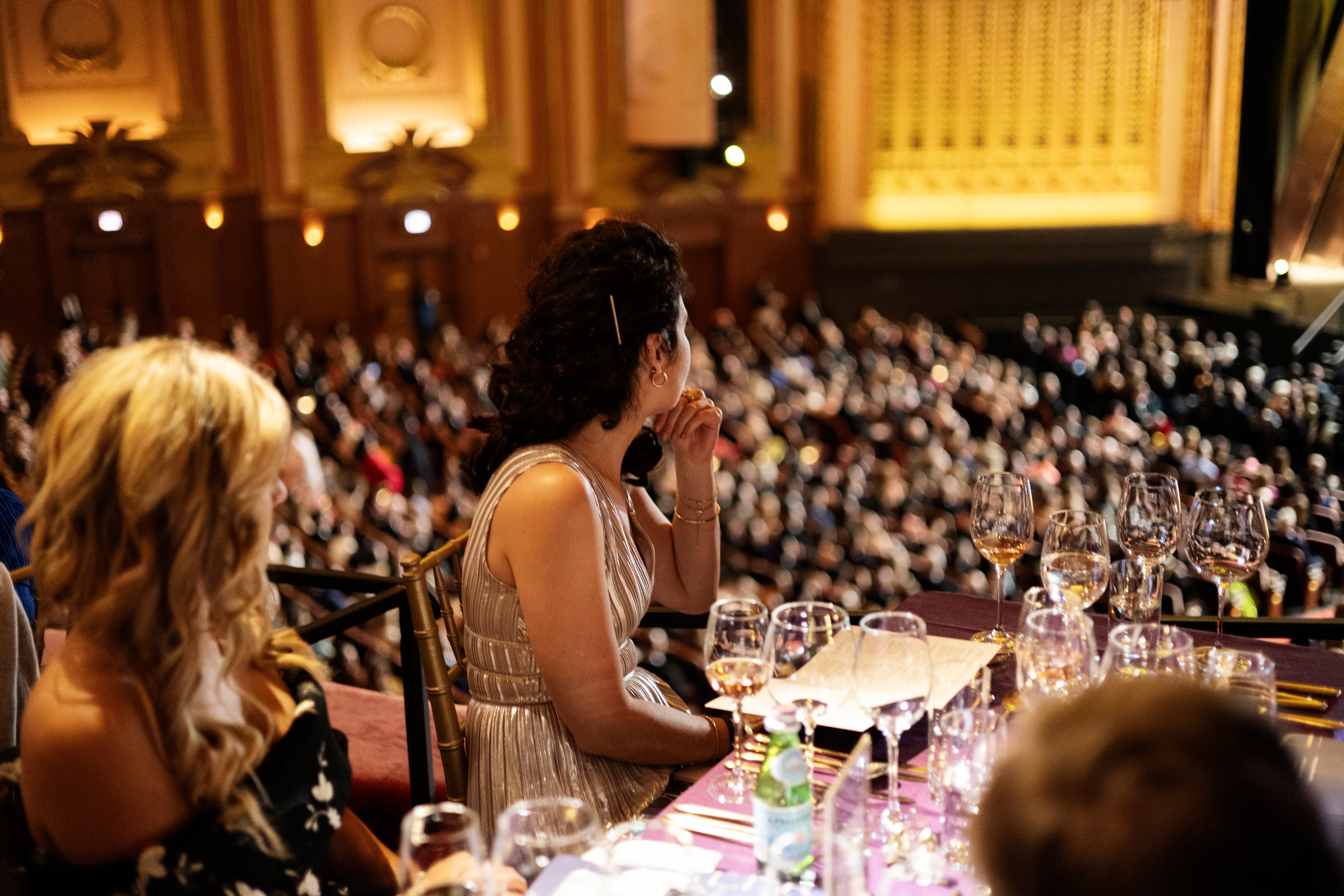 A woman seated at a formal table in a theater, overlooking a crowded award event.