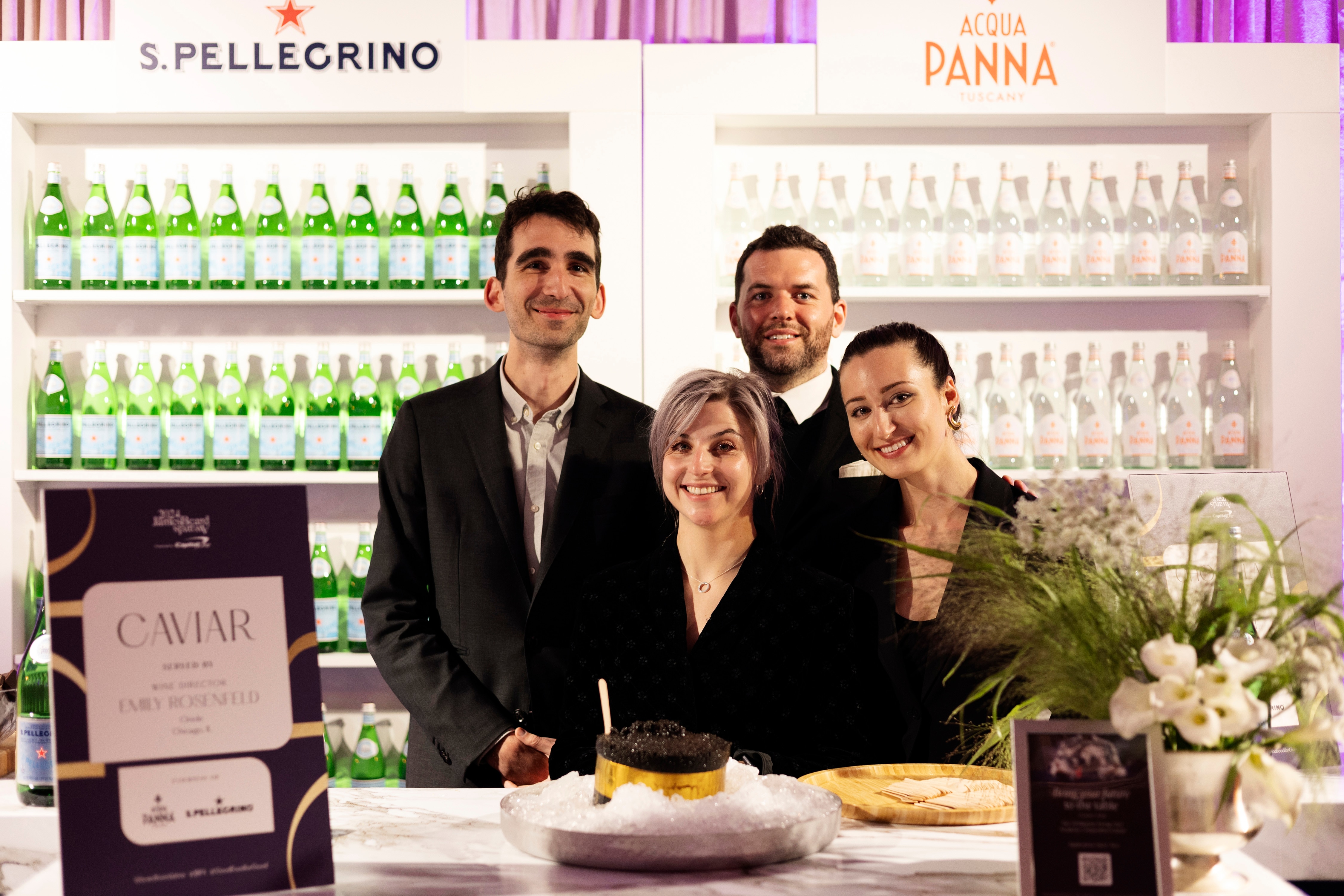 Four people standing at a bar with S.Pellegrino and Acqua Panna bottles displayed behind them.