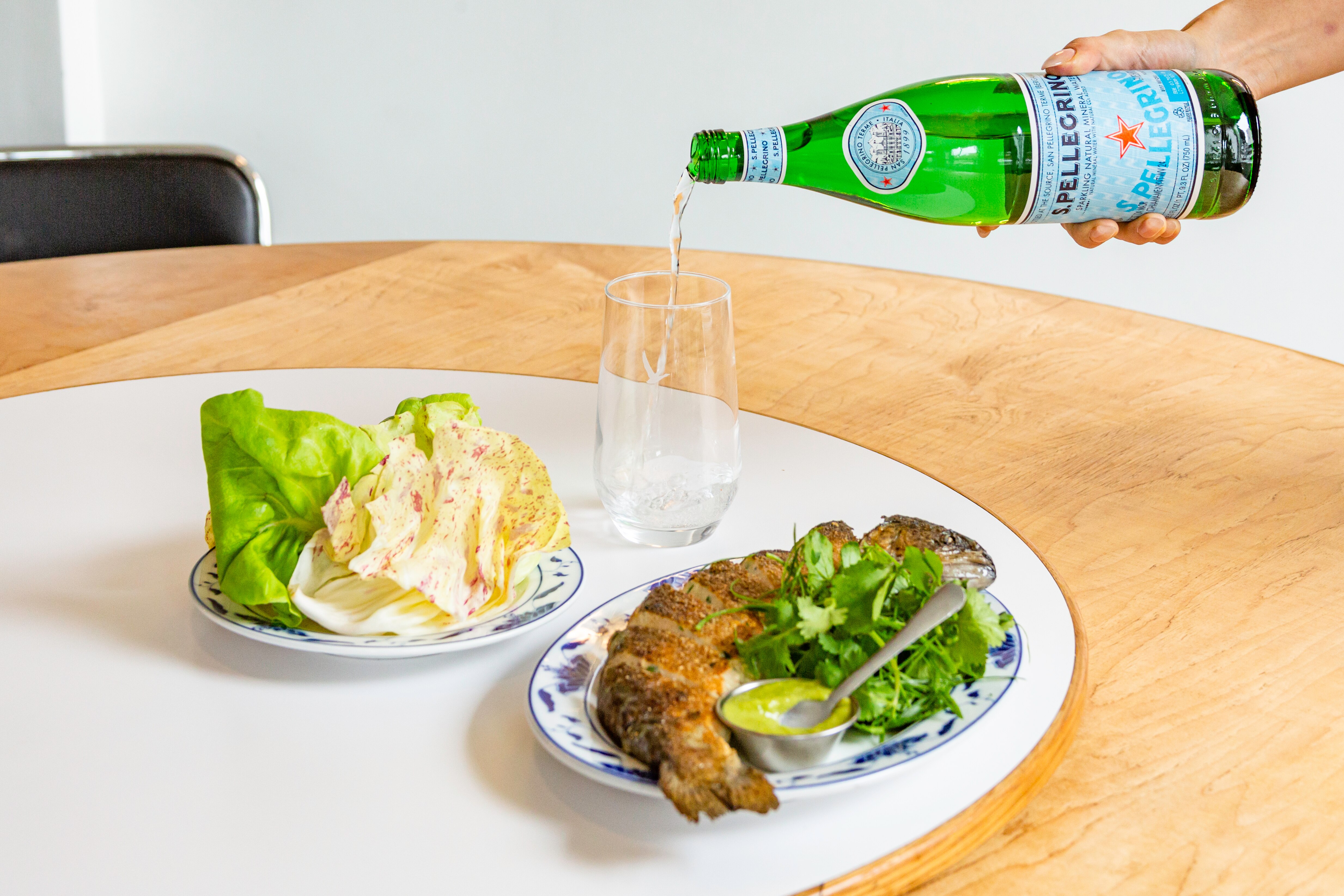 S.Pellegrino bottle being poured into a glass alongside a plate of fish and salad on a wooden table.