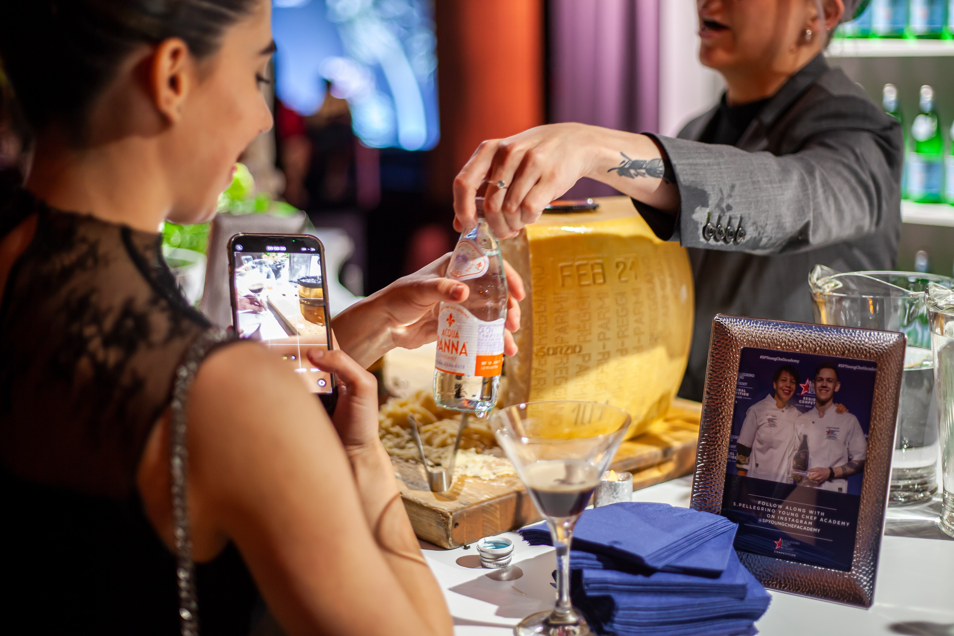 A woman takes a photo of a chef holding an Acqua Panna bottle near a large wheel of cheese.