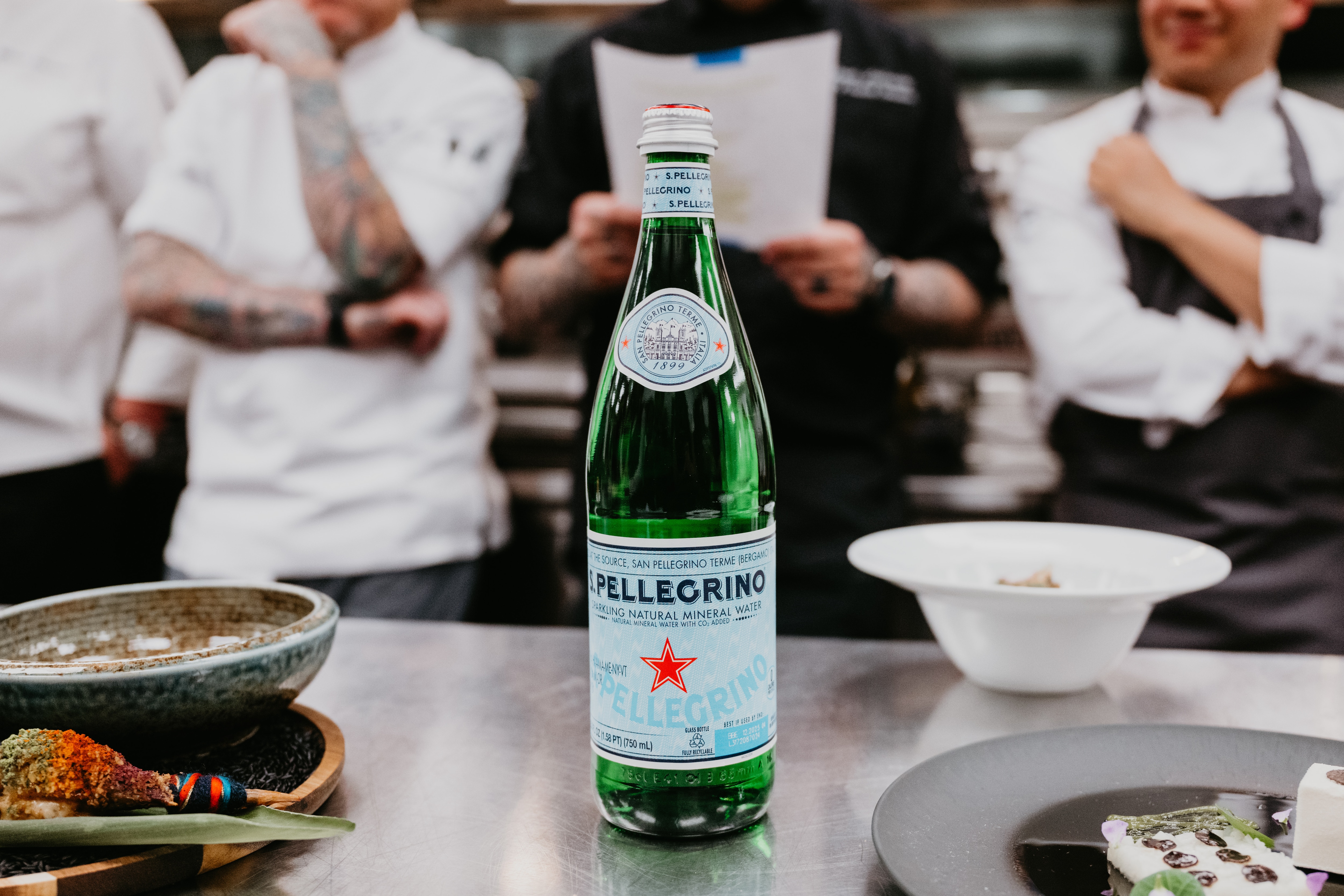 A bottle of S.Pellegrino sits before chefs in a professional kitchen