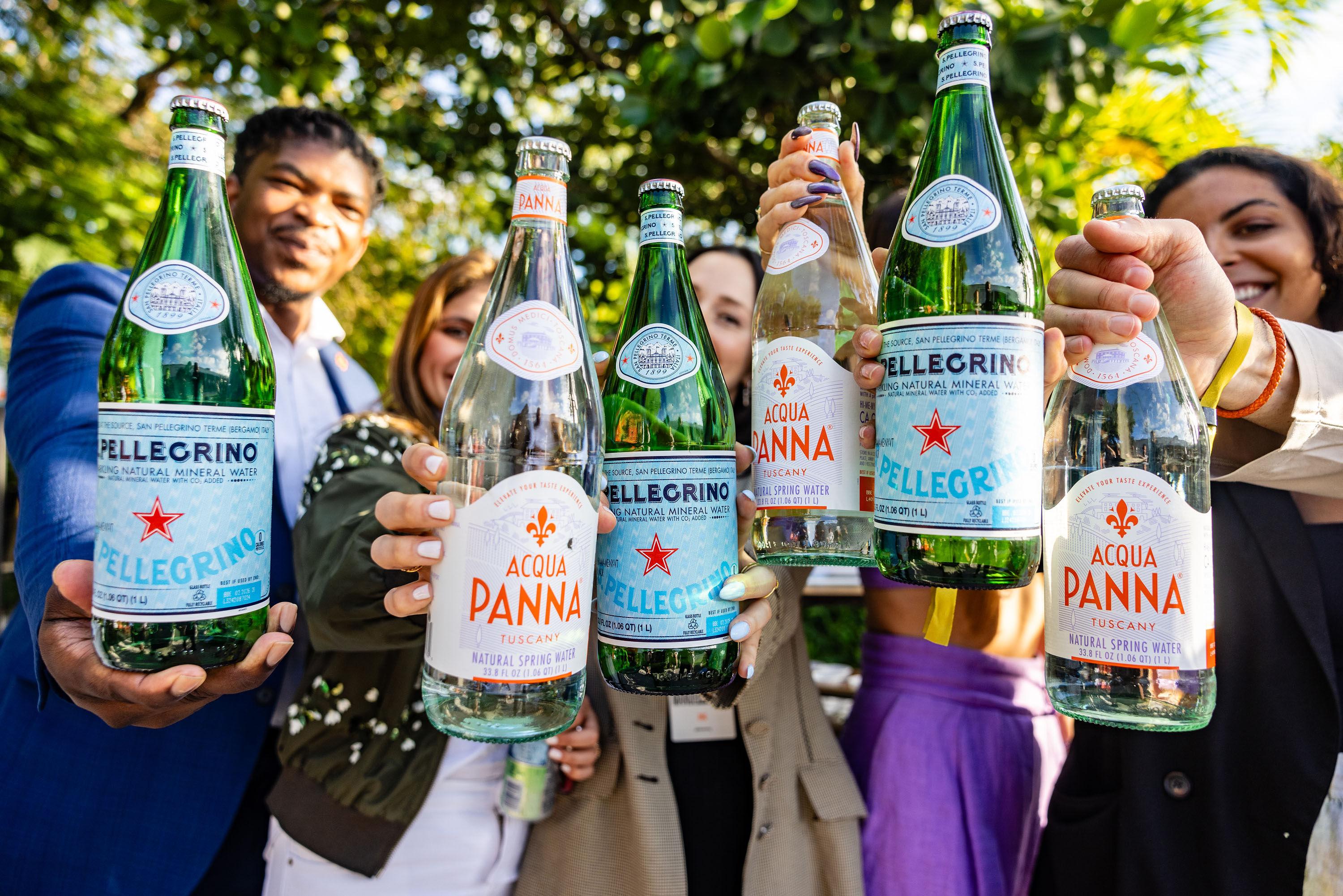 Group holding bottles of S.Pellegrino and Acqua Panna, smiling towards the camera outdoors.