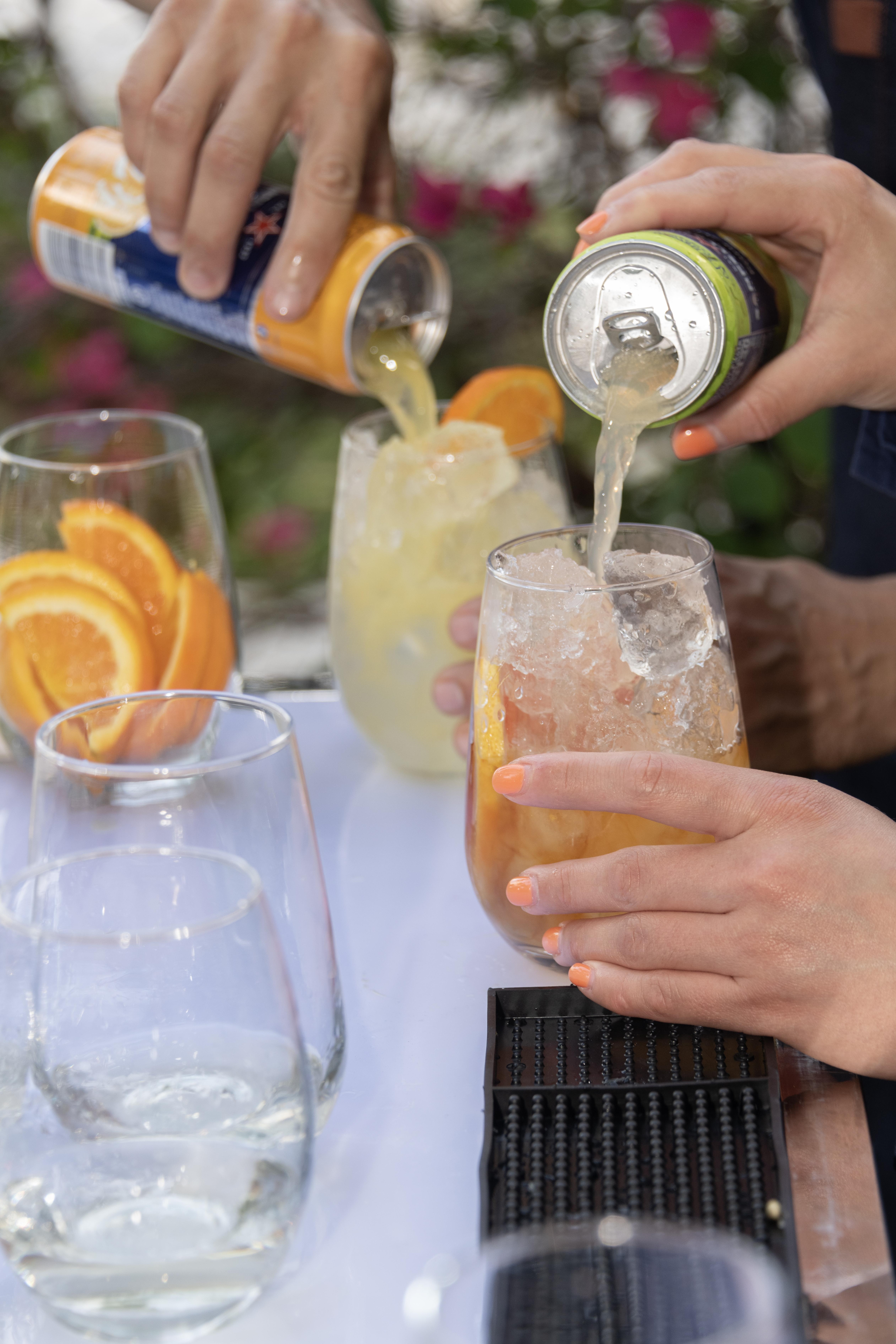 Hands pouring drinks from cans into glasses filled with ice and orange slices.