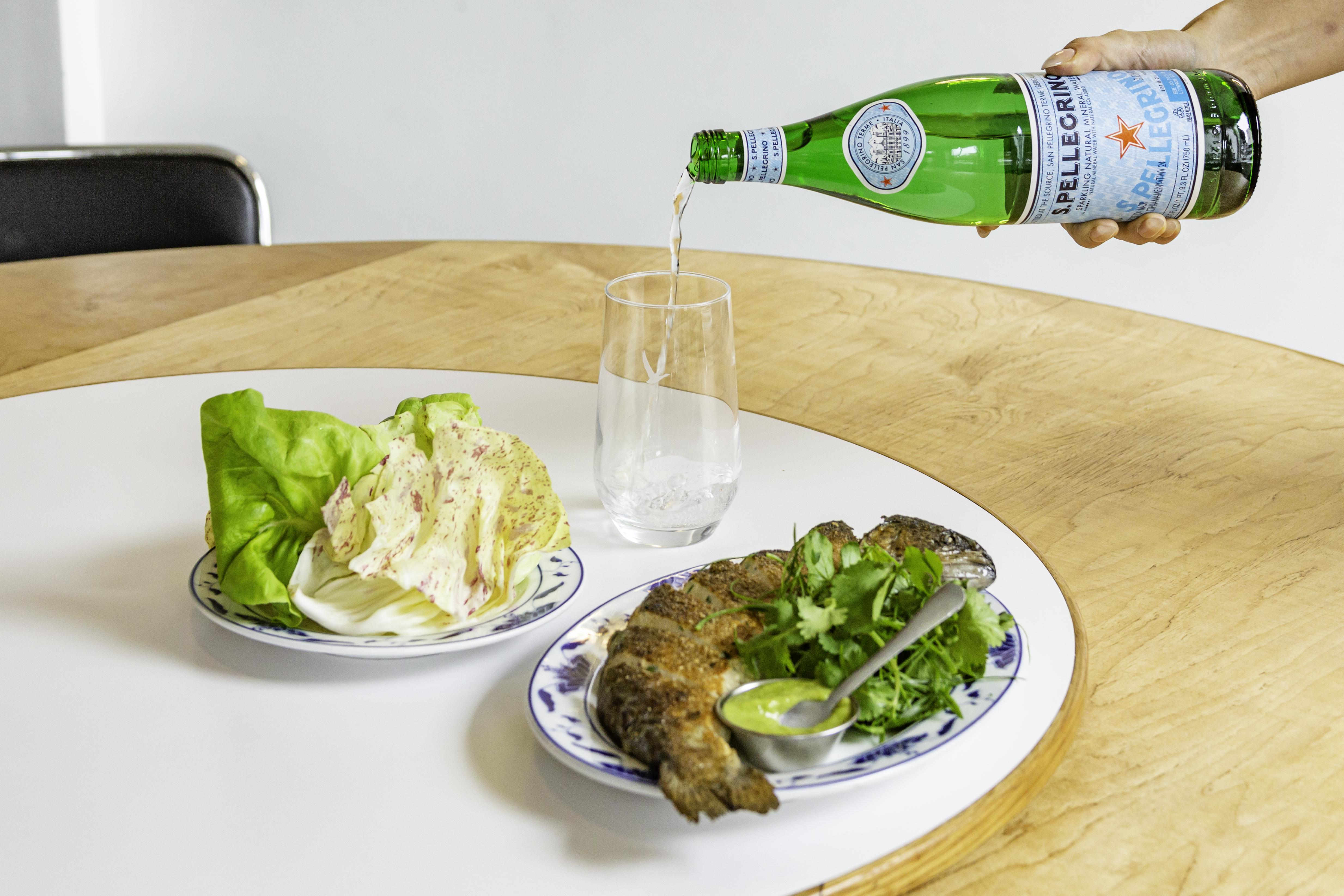 S.Pellegrino bottle being poured into a glass alongside a plate of fish and salad on a wooden table.